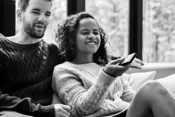 black & white photograph of a couple watching a movie. The woman is leaning back into the man and is pointing a remote control at the tv.