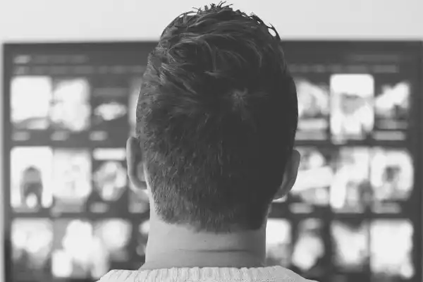 A black and white photo taken from behind of a man's head and back as he sits looking at a tv screen.